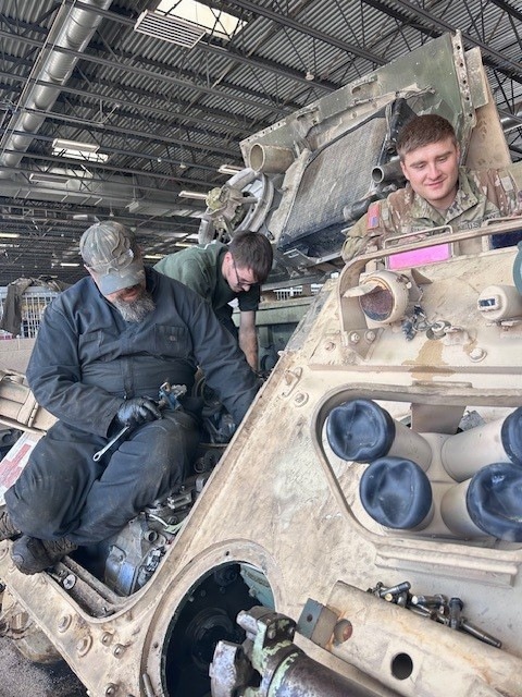 Three individuals are performing maintenance or repair work on a military armored vehicle inside a large indoor facility, likely a maintenance bay or depot. Two workers in civilian coveralls are working on the vehicle's hull components, while a soldier in camouflage uniform observes or assists from an open hatch on top of the vehicle. The vehicle appears to be a desert-tan painted armored fighting vehicle with visible wear and disassembled components.