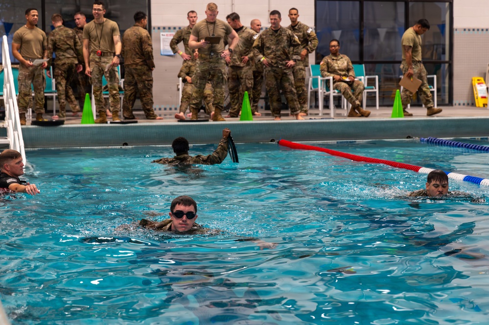 Military personnel are conducting a water survival or swim qualification training exercise in an indoor pool facility. Several soldiers in full camouflage uniforms are swimming in the pool while a larger group of uniformed observers watches from the pool deck. One swimmer appears to be holding equipment above the water with a raised fist, suggesting a specific swim technique or equipment carry drill.