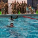 Military personnel are conducting a water survival or swim qualification training exercise in an indoor pool facility. Several soldiers in full camouflage uniforms are swimming in the pool while a larger group of uniformed observers watches from the pool deck. One swimmer appears to be holding equipment above the water with a raised fist, suggesting a specific swim technique or equipment carry drill.