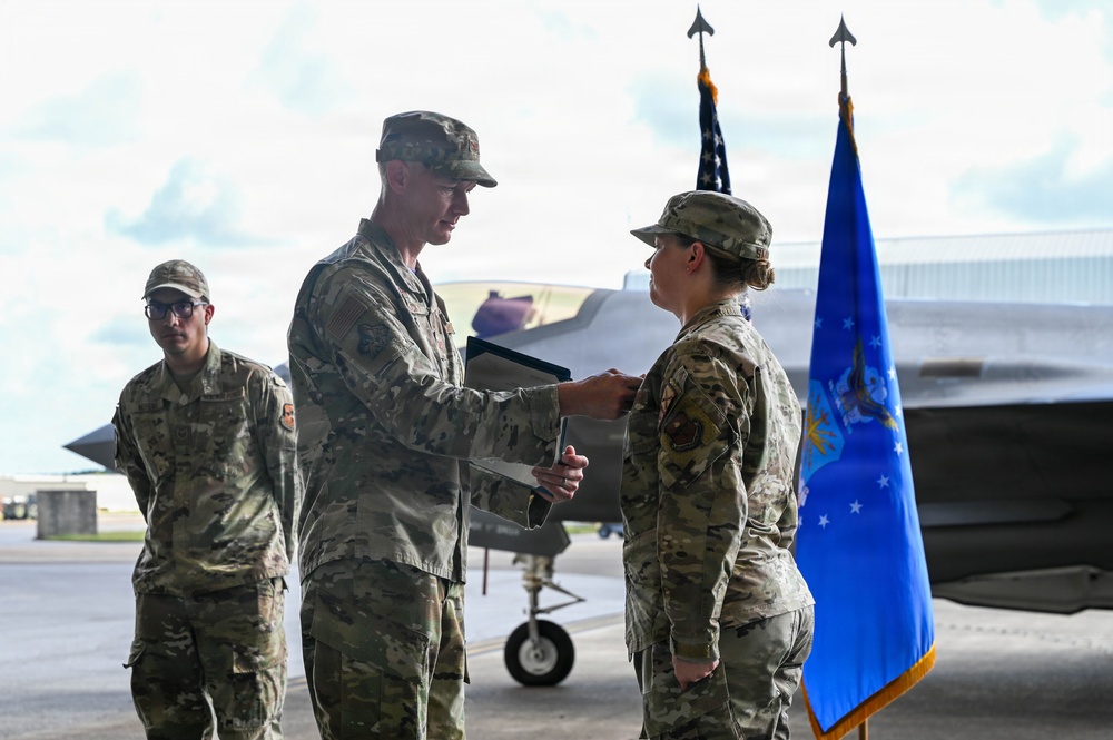 A military ceremony is taking place on an aircraft flight line, where a senior officer appears to be presenting an award or document to a female service member. A blue Air Force flag and American flag are visible in the background, along with what appears to be an F-35 fighter jet. A third service member stands in the background observing the ceremony.