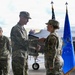 A military ceremony is taking place on an aircraft flight line, where a senior officer appears to be presenting an award or document to a female service member. A blue Air Force flag and American flag are visible in the background, along with what appears to be an F-35 fighter jet. A third service member stands in the background observing the ceremony.
