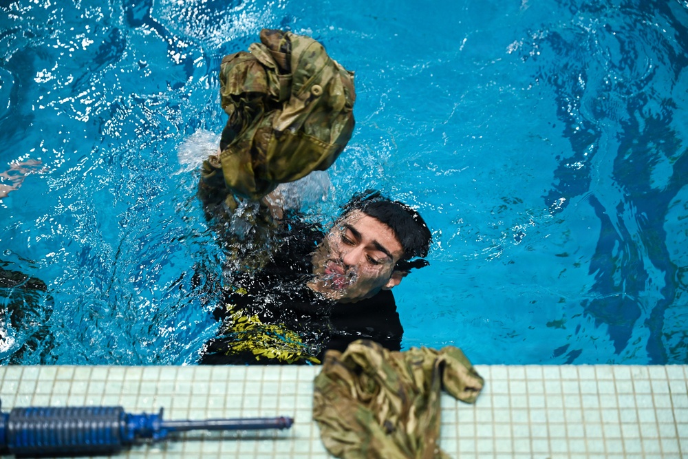 A person in a swimming pool is removing military camouflage clothing while in the water, holding a wet uniform jacket aloft. Additional camouflage garments and what appears to be a blue training rifle are visible on the pool deck. This appears to be a military water survival or combat swimming training exercise.