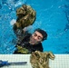 A person in a swimming pool is removing military camouflage clothing while in the water, holding a wet uniform jacket aloft. Additional camouflage garments and what appears to be a blue training rifle are visible on the pool deck. This appears to be a military water survival or combat swimming training exercise.