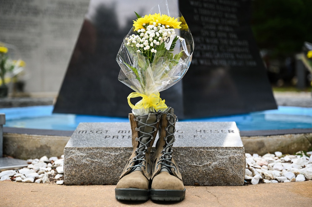 A memorial tribute featuring a pair of military combat boots with a bouquet of yellow and white flowers placed inside them, positioned in front of a granite memorial stone. The stone bears an inscription referencing 'MSGT MI...G HEISER' and 'PATR...B FL', indicating a military memorial site. The arrangement is a traditional 'battlefield cross' variant honoring a fallen service member.