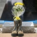 A memorial tribute featuring a pair of military combat boots with a bouquet of yellow and white flowers placed inside them, positioned in front of a granite memorial stone. The stone bears an inscription referencing 'MSGT MI...G HEISER' and 'PATR...B FL', indicating a military memorial site. The arrangement is a traditional 'battlefield cross' variant honoring a fallen service member.