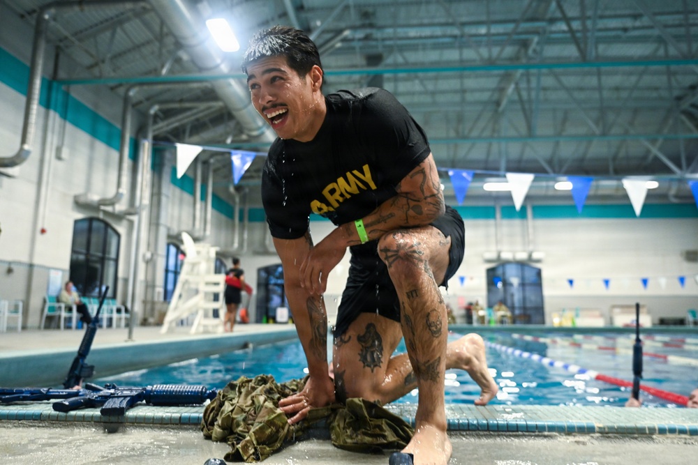 A heavily tattooed man wearing a black U.S. Army t-shirt is crouching at the edge of an indoor swimming pool, soaking wet and laughing. He appears to have just exited the pool during what looks like a military swim training or competition event. Replica training rifles and military gear are visible on the pool deck beside him.