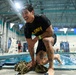 A heavily tattooed man wearing a black U.S. Army t-shirt is crouching at the edge of an indoor swimming pool, soaking wet and laughing. He appears to have just exited the pool during what looks like a military swim training or competition event. Replica training rifles and military gear are visible on the pool deck beside him.