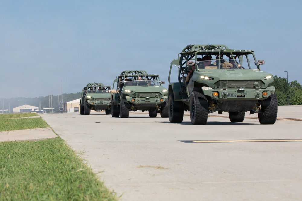 A convoy of three military light tactical vehicles, appearing to be Infantry Squad Vehicles (ISVs) or similar open-frame reconnaissance vehicles, travel in formation along a paved military airfield or road. The vehicles are painted in olive drab/military green and are occupied by personnel in military attire. The setting appears to be a U.S. military installation during daytime with clear weather conditions.