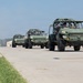 A convoy of three military light tactical vehicles, appearing to be Infantry Squad Vehicles (ISVs) or similar open-frame reconnaissance vehicles, travel in formation along a paved military airfield or road. The vehicles are painted in olive drab/military green and are occupied by personnel in military attire. The setting appears to be a U.S. military installation during daytime with clear weather conditions.