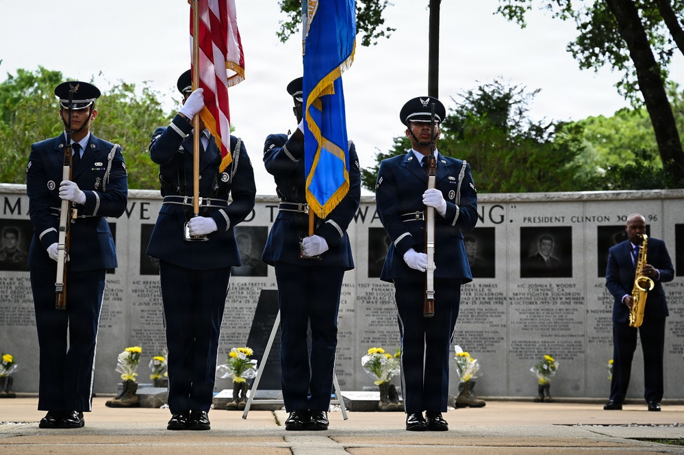 A U.S. Air Force color guard stands at attention during a memorial ceremony, holding the American flag and an Air Force flag in front of a memorial wall. The wall features engraved names, portraits, and a quote attributed to President Clinton dated 1996, suggesting this is a commemoration related to a specific military tragedy. Yellow flowers are placed at the base of the memorial, and a musician playing a saxophone stands to the right.
