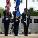 A U.S. Air Force color guard stands at attention during a memorial ceremony, holding the American flag and an Air Force flag in front of a memorial wall. The wall features engraved names, portraits, and a quote attributed to President Clinton dated 1996, suggesting this is a commemoration related to a specific military tragedy. Yellow flowers are placed at the base of the memorial, and a musician playing a saxophone stands to the right.
