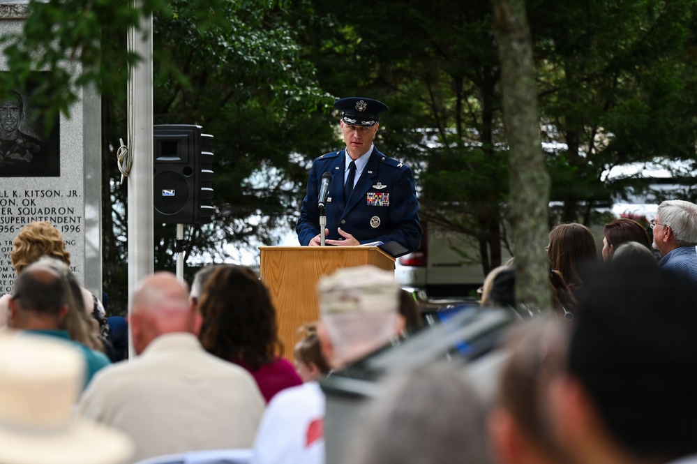 A United States Air Force officer in dress blue uniform is delivering a speech at a podium during what appears to be a military memorial or commemorative ceremony held outdoors. A crowd of civilians and attendees is seated facing the speaker, with a stone memorial monument visible to the left bearing inscribed text and a portrait relief. The setting is surrounded by trees, suggesting a park or memorial grounds.