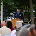 A United States Air Force officer in dress blue uniform is delivering a speech at a podium during what appears to be a military memorial or commemorative ceremony held outdoors. A crowd of civilians and attendees is seated facing the speaker, with a stone memorial monument visible to the left bearing inscribed text and a portrait relief. The setting is surrounded by trees, suggesting a park or memorial grounds.