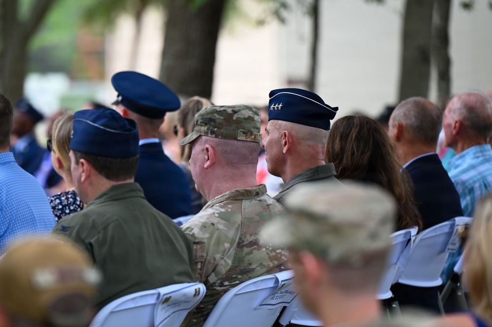 A group of military personnel and civilians are seated outdoors at what appears to be a formal military ceremony or event. Several uniformed individuals are visible, including Air Force officers wearing service dress caps and at least one soldier in Army Combat Uniform (ACU/OCP camouflage). The image is taken from behind the seated audience, showing the backs of attendees.