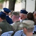 A group of military personnel and civilians are seated outdoors at what appears to be a formal military ceremony or event. Several uniformed individuals are visible, including Air Force officers wearing service dress caps and at least one soldier in Army Combat Uniform (ACU/OCP camouflage). The image is taken from behind the seated audience, showing the backs of attendees.