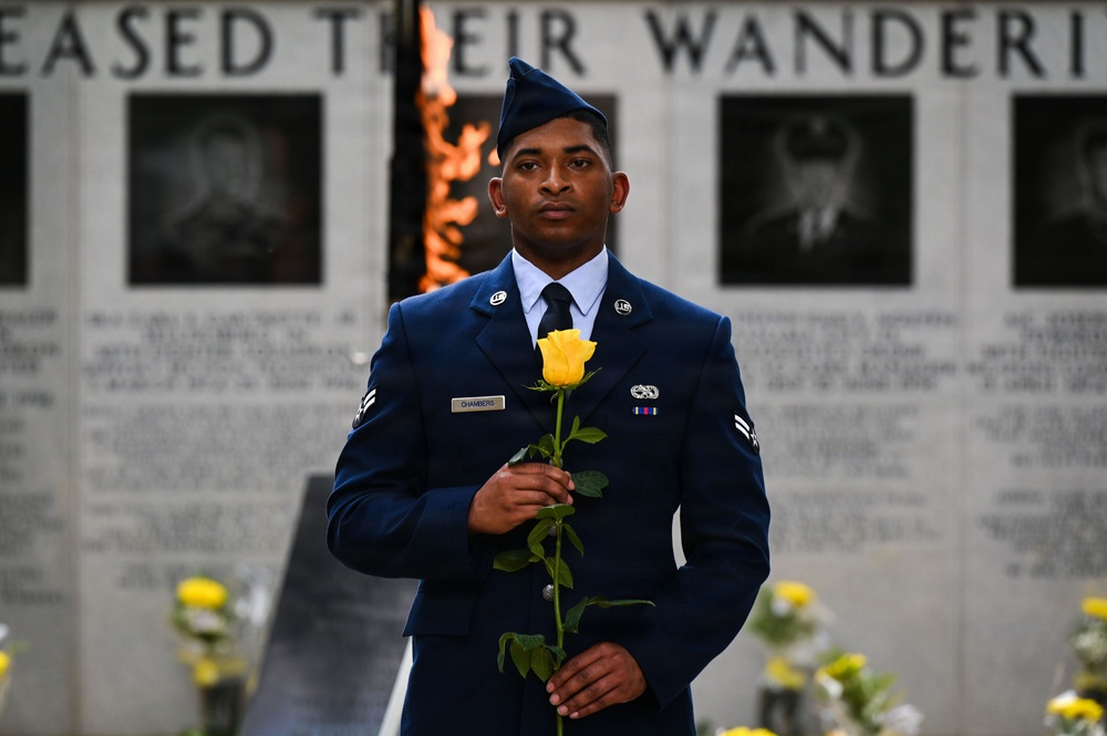 A U.S. Air Force servicemember in dress blue uniform stands solemnly at what appears to be a memorial wall, holding a single yellow rose. The background features engraved names and portrait reliefs on a stone memorial wall, along with an eternal flame visible behind the subject. The scene suggests a formal memorial ceremony or tribute.