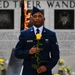 A U.S. Air Force servicemember in dress blue uniform stands solemnly at what appears to be a memorial wall, holding a single yellow rose. The background features engraved names and portrait reliefs on a stone memorial wall, along with an eternal flame visible behind the subject. The scene suggests a formal memorial ceremony or tribute.