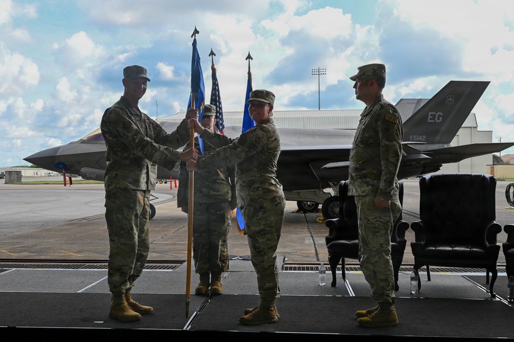 A military change of command ceremony is taking place on an outdoor stage at an air base. A guidon (unit flag) is being passed between personnel in OCP (Operational Camouflage Pattern) uniforms, signifying the transfer of command authority. An F-35 fighter jet bearing the tail code 'EG' and serial number '5142' is visible in the background, indicating this is likely Eglin Air Force Base.