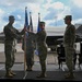 A military change of command ceremony is taking place on an outdoor stage at an air base. A guidon (unit flag) is being passed between personnel in OCP (Operational Camouflage Pattern) uniforms, signifying the transfer of command authority. An F-35 fighter jet bearing the tail code 'EG' and serial number '5142' is visible in the background, indicating this is likely Eglin Air Force Base.