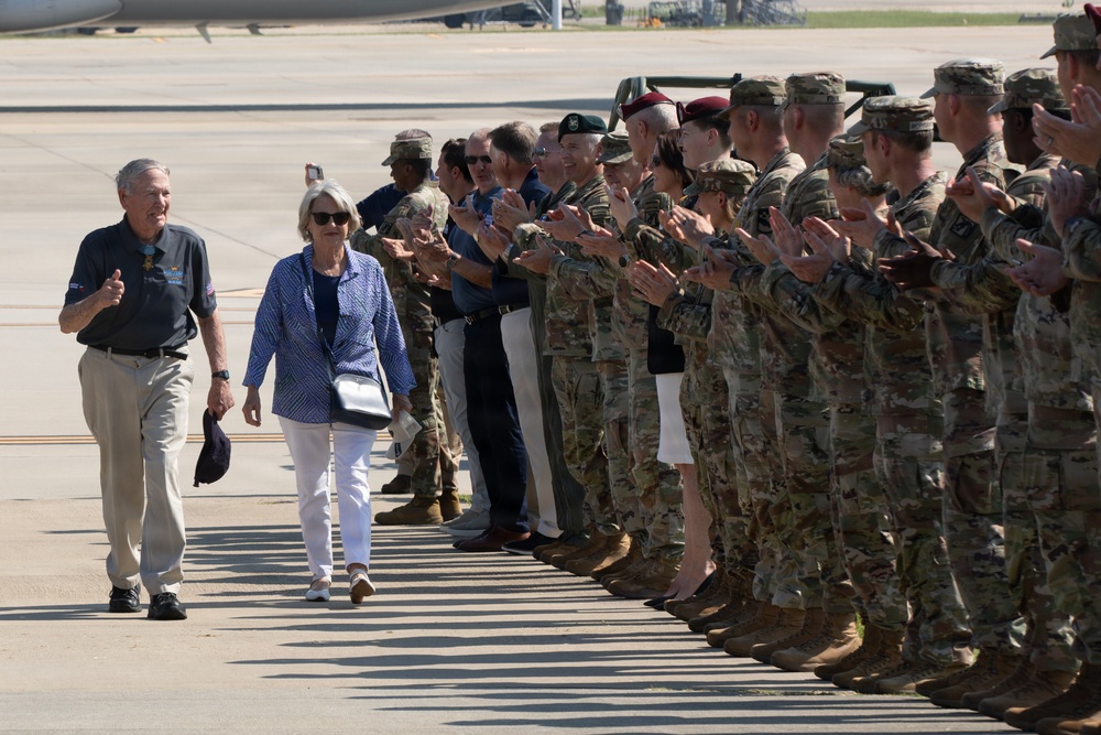 Two civilians, an older man and woman, walk along a tarmac while a long line of military personnel in camouflage uniforms applaud them. The man appears to be wearing a medal around his neck, suggesting this is an official military honor ceremony. The setting appears to be a military airfield or base.