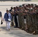 Two civilians, an older man and woman, walk along a tarmac while a long line of military personnel in camouflage uniforms applaud them. The man appears to be wearing a medal around his neck, suggesting this is an official military honor ceremony. The setting appears to be a military airfield or base.