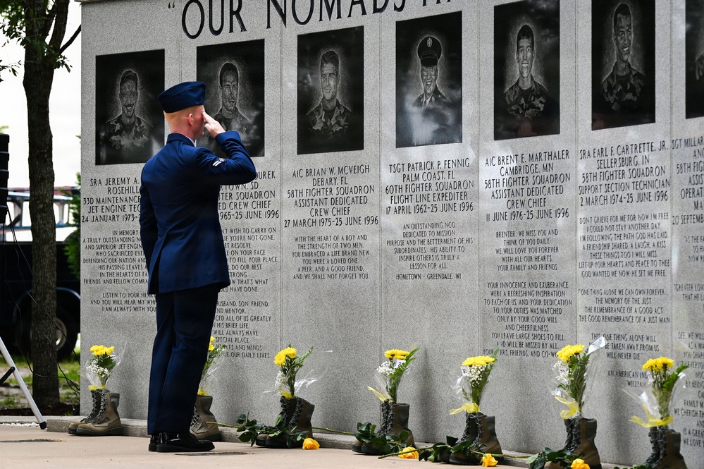A uniformed U.S. Air Force service member renders a salute at a memorial wall honoring fallen airmen, primarily from the 58th Fighter Squadron who died on 25 June 1996. The granite memorial wall features engraved portraits and biographical inscriptions of the deceased, with military boots and yellow flowers placed at its base as tributes. The header text reads 'OUR NOMADS' and the setting appears to be an outdoor memorial ceremony.