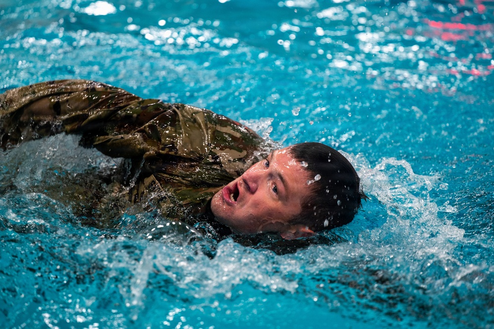 A person wearing military camouflage uniform is swimming in an indoor pool, with their head raised above the water. The individual appears to be undergoing military water survival or combat swimmer training, as evidenced by the fully clothed swim in what appears to be a controlled pool environment. The image captures intense physical exertion with significant water splashing around the swimmer.