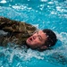 A person wearing military camouflage uniform is swimming in an indoor pool, with their head raised above the water. The individual appears to be undergoing military water survival or combat swimmer training, as evidenced by the fully clothed swim in what appears to be a controlled pool environment. The image captures intense physical exertion with significant water splashing around the swimmer.