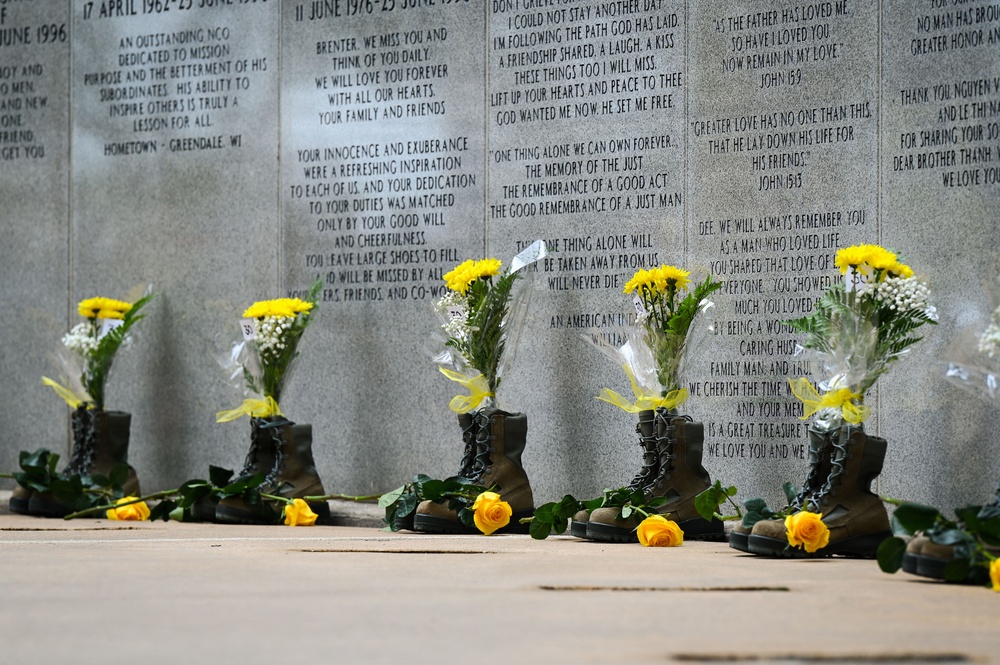 A military memorial wall with engraved tributes and inscriptions dedicated to fallen soldiers. Several pairs of military combat boots have been placed at the base of the wall, each filled with yellow flowers (sunflowers, roses, and baby's breath). Yellow roses are also laid flat on the ground in front of the boots, creating a solemn and heartfelt tribute.
