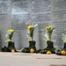 A military memorial wall with engraved tributes and inscriptions dedicated to fallen soldiers. Several pairs of military combat boots have been placed at the base of the wall, each filled with yellow flowers (sunflowers, roses, and baby's breath). Yellow roses are also laid flat on the ground in front of the boots, creating a solemn and heartfelt tribute.