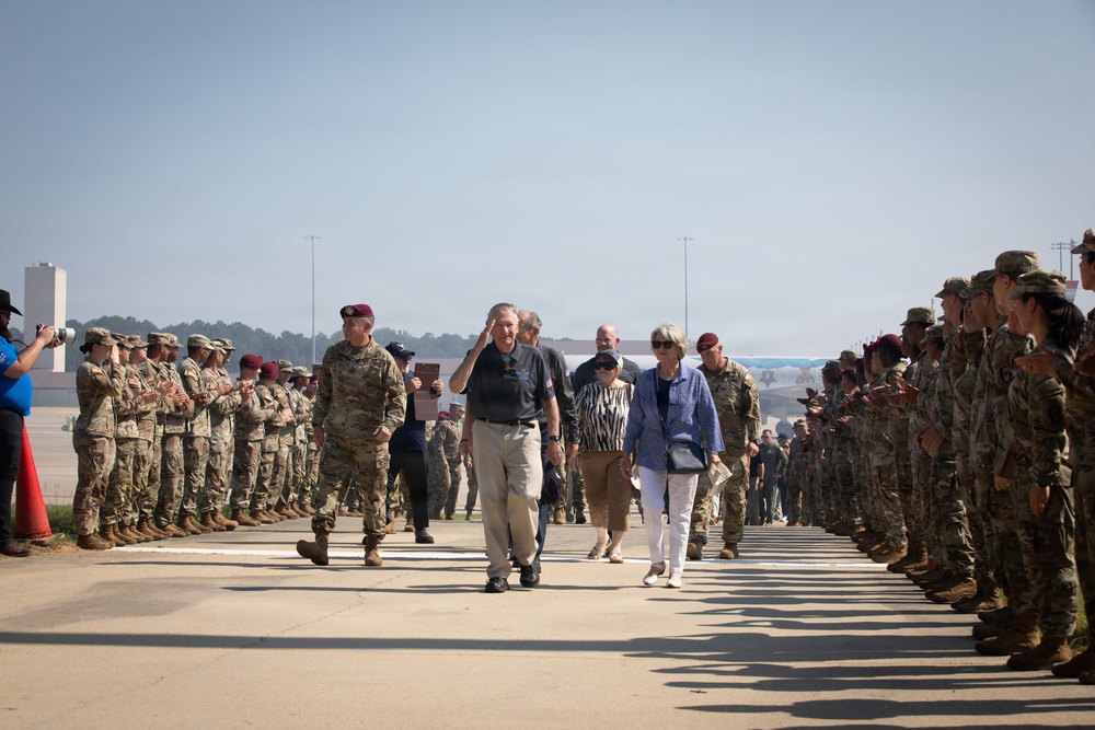 A military honors ceremony is taking place on what appears to be a military installation tarmac or parade ground. A civilian dignitary and military personnel are walking through a corridor formed by two lines of uniformed soldiers standing at attention. The soldiers are wearing camouflage uniforms and some wear distinctive maroon/red berets, suggesting a paratrooper or airborne unit.