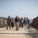 A military honors ceremony is taking place on what appears to be a military installation tarmac or parade ground. A civilian dignitary and military personnel are walking through a corridor formed by two lines of uniformed soldiers standing at attention. The soldiers are wearing camouflage uniforms and some wear distinctive maroon/red berets, suggesting a paratrooper or airborne unit.