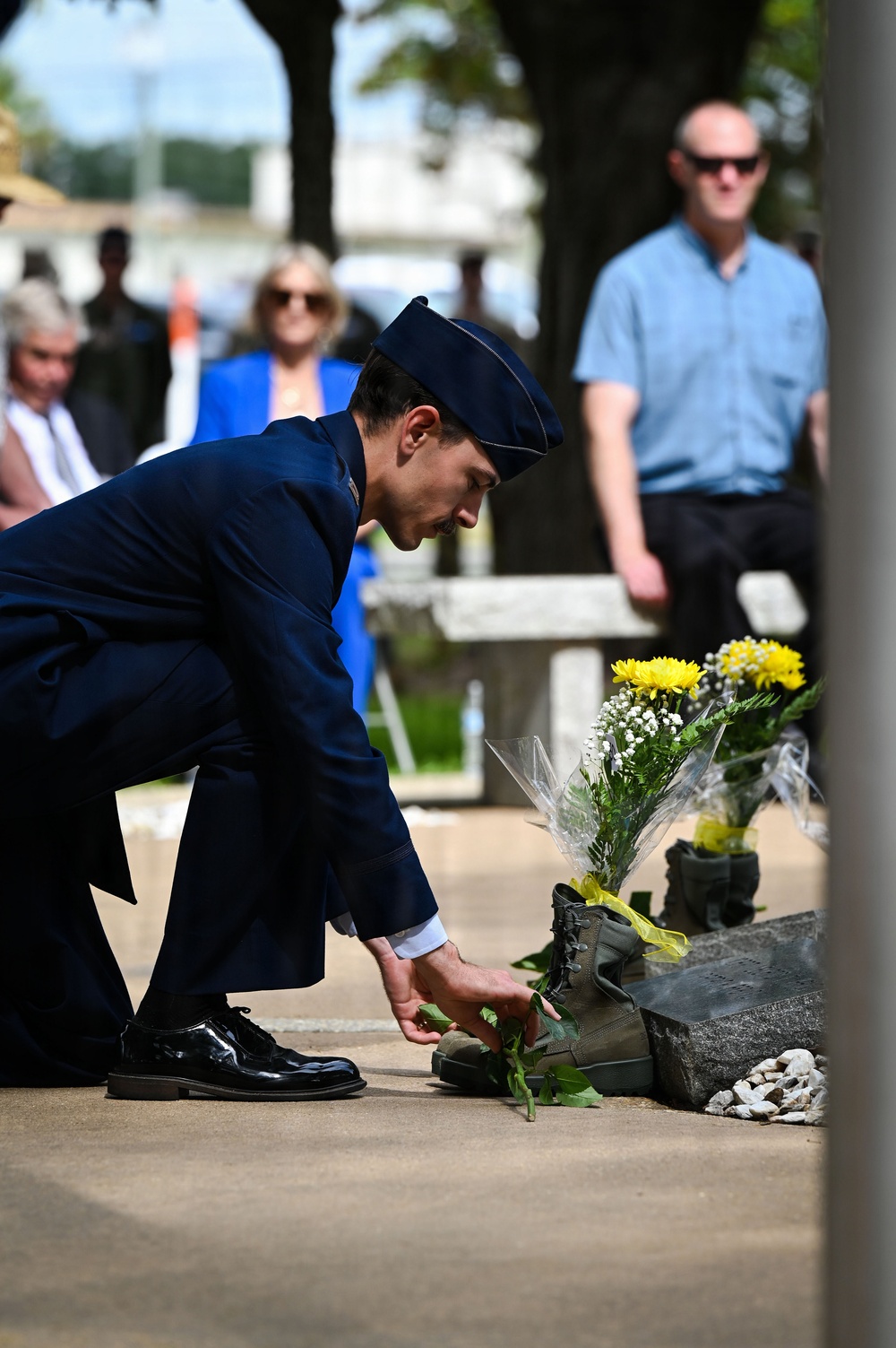 A uniformed military service member, wearing a dark blue dress uniform and service cap consistent with the U.S. Air Force, bends down to place a flower or greenery at a memorial site. The memorial features a combat boot, yellow flowers, and a stone or granite base, suggesting a fallen soldier tribute. Civilian onlookers are visible in the blurred background, indicating a formal commemorative ceremony.