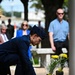 A uniformed military service member, wearing a dark blue dress uniform and service cap consistent with the U.S. Air Force, bends down to place a flower or greenery at a memorial site. The memorial features a combat boot, yellow flowers, and a stone or granite base, suggesting a fallen soldier tribute. Civilian onlookers are visible in the blurred background, indicating a formal commemorative ceremony.