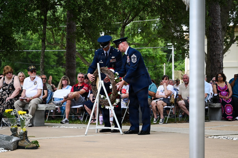 Two U.S. Air Force personnel in dress blue uniforms are placing a floral wreath on a white stand during what appears to be a memorial ceremony. A seated audience of civilians and veterans watches the solemn event, which takes place on an outdoor plaza surrounded by trees. Combat boots with flowers placed in them are visible in the foreground, a common tribute symbol at military memorial services.