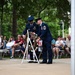 Two U.S. Air Force personnel in dress blue uniforms are placing a floral wreath on a white stand during what appears to be a memorial ceremony. A seated audience of civilians and veterans watches the solemn event, which takes place on an outdoor plaza surrounded by trees. Combat boots with flowers placed in them are visible in the foreground, a common tribute symbol at military memorial services.