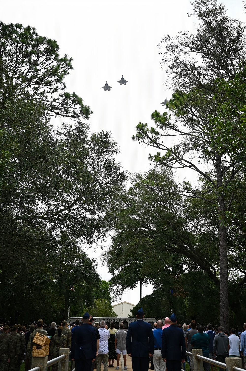 A military flyover ceremony is taking place, with three jet aircraft flying in a missing man formation over a tree-lined pathway. A crowd of military personnel in uniform and civilians are gathered below, looking skyward toward the aircraft. A partially visible inscription on a structure in the background reads '...DS HERE CEASED THEIR WANDER...', suggesting a memorial or cemetery setting.