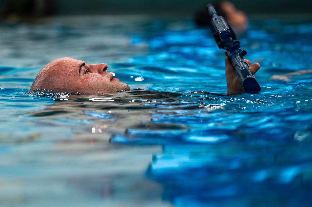 A bald male individual is swimming on his back in a pool while holding a rifle or training weapon above the water surface with one arm extended upward. The weapon appears to be a blue/dark-colored training firearm or replica. This appears to be a military or law enforcement water survival training exercise.