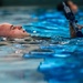 A bald male individual is swimming on his back in a pool while holding a rifle or training weapon above the water surface with one arm extended upward. The weapon appears to be a blue/dark-colored training firearm or replica. This appears to be a military or law enforcement water survival training exercise.