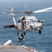 A U.S. Navy MH-60S Seahawk helicopter (bureau number/side number '62') hovers over the deck of a naval vessel at sea. A rescue/hoist operation is underway, with at least one person being lowered or raised via a hoist cable. The ocean background and ship deck confirm an open-water maritime environment.