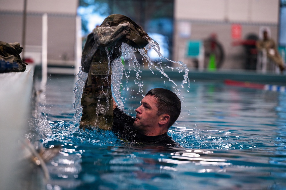 A male individual in military uniform is submerged in an indoor swimming pool, lifting soaking wet camouflage clothing above the water surface. Water is visibly cascading off the garment, suggesting a military water survival or combat swimming training exercise. Additional personnel and pool safety equipment are visible in the background.