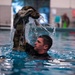 A male individual in military uniform is submerged in an indoor swimming pool, lifting soaking wet camouflage clothing above the water surface. Water is visibly cascading off the garment, suggesting a military water survival or combat swimming training exercise. Additional personnel and pool safety equipment are visible in the background.