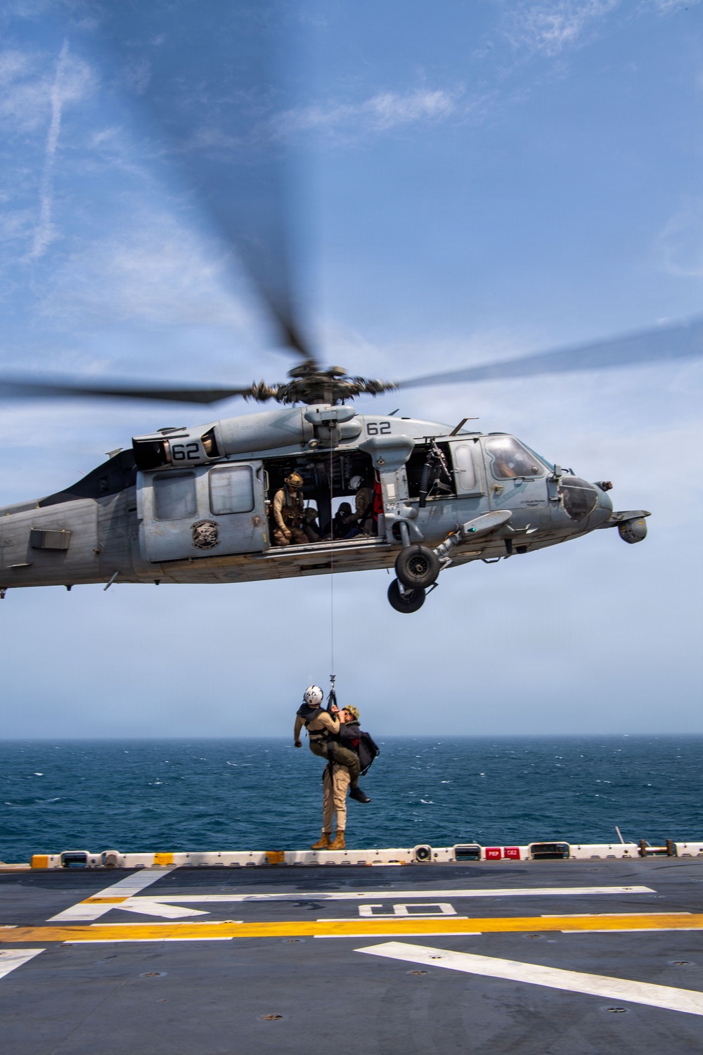 A military helicopter numbered '62' hovers over a naval vessel's flight deck while conducting a hoist rescue or training operation. A rescue swimmer or aircrew member is suspended on a hoist cable, holding a second person in a rescue carry position. The scene appears to be a search and rescue (SAR) training exercise conducted at sea.