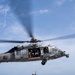 A military helicopter numbered '62' hovers over a naval vessel's flight deck while conducting a hoist rescue or training operation. A rescue swimmer or aircrew member is suspended on a hoist cable, holding a second person in a rescue carry position. The scene appears to be a search and rescue (SAR) training exercise conducted at sea.
