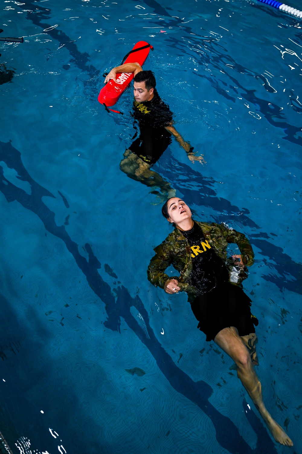 Two individuals in military-style uniforms are in an indoor swimming pool during what appears to be a water rescue training exercise. One person, positioned higher in the frame, is holding a red lifeguard rescue buoy above the water while swimming. The second person is floating on their back in a simulated rescue victim position, wearing camouflage and a black shirt with yellow lettering.