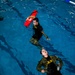 Two individuals in military-style uniforms are in an indoor swimming pool during what appears to be a water rescue training exercise. One person, positioned higher in the frame, is holding a red lifeguard rescue buoy above the water while swimming. The second person is floating on their back in a simulated rescue victim position, wearing camouflage and a black shirt with yellow lettering.