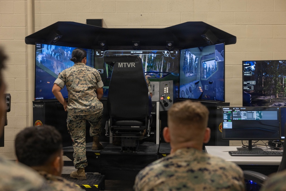 Military personnel in camouflage uniforms are gathered around an MTVR (Medium Tactical Vehicle Replacement) driving simulator in what appears to be a training facility. The simulator features a multi-screen wraparound display showing a forested environment, with a central operator seat labeled 'MTVR'. A separate instructor/monitoring station with additional screens is visible to the right.