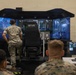 Military personnel in camouflage uniforms are gathered around an MTVR (Medium Tactical Vehicle Replacement) driving simulator in what appears to be a training facility. The simulator features a multi-screen wraparound display showing a forested environment, with a central operator seat labeled 'MTVR'. A separate instructor/monitoring station with additional screens is visible to the right.