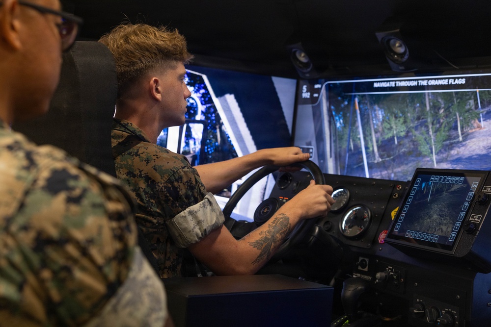 A young person in military camouflage uniform is operating a vehicle driving simulator, gripping a steering wheel while viewing a large screen displaying a forested off-road environment. A second individual in military camouflage is visible in the foreground observing. The simulator appears to be a military-grade training system with multiple displays and control panels.