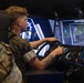A young person in military camouflage uniform is operating a vehicle driving simulator, gripping a steering wheel while viewing a large screen displaying a forested off-road environment. A second individual in military camouflage is visible in the foreground observing. The simulator appears to be a military-grade training system with multiple displays and control panels.